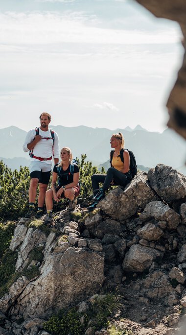 Drei Wanderer auf einem felsigen Bergkamm mit Blick auf die Berge im Hintergrund, eine Frau sitzt auf einem Felsen, die anderen beiden stehen. | © Kleinwalsertal Tourismus eGen • Fotograf: Stefan Klauser 
