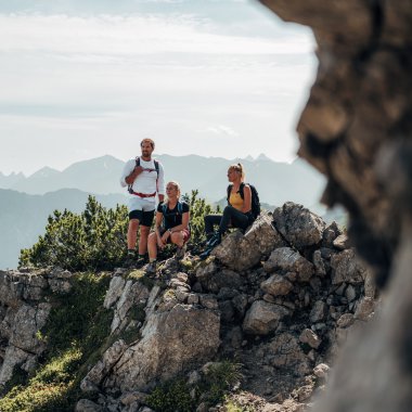 Drei Wanderer auf einem felsigen Bergkamm mit Blick auf die Berge im Hintergrund, eine Frau sitzt auf einem Felsen, die anderen beiden stehen. | © Kleinwalsertal Tourismus eGen • Fotograf: Stefan Klauser 