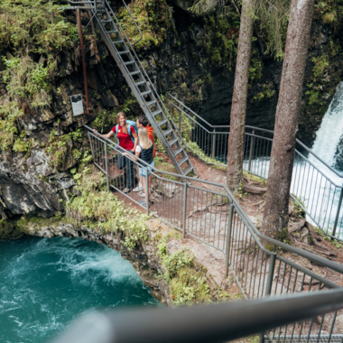 Zwei Personen in roter Kleidung auf einem Holzsteg im Wald neben einem Wasserfall, umgeben von grünen Bäumen. | ©  Kleinwalsertal Tourismus eGen • Fotograf: Stefan Klauser 