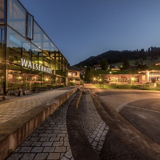 © Kleinwalsertal Tourismus eGen • Fotograf: Steffen Berschin Abendaufnahme des Walserhauses mit reflektierenden Glasfenstern, umgeben von Bergen und beleuchteter Landschaft. | © Kleinwalsertal Tourismus eGen • Fotograf: Steffen Berschin