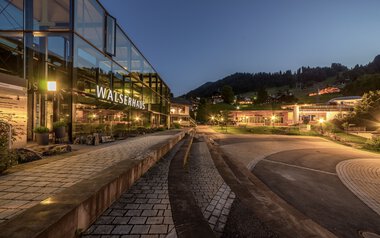 Abendaufnahme des Walserhauses mit reflektierenden Glasfenstern, umgeben von Bergen und beleuchteter Landschaft. | © Kleinwalsertal Tourismus eGen • Fotograf: Steffen Berschin 