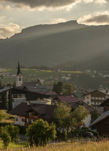 Bergdorf mit Kirche und umliegenden Bergen bei Sonnenaufgang oder -untergang, warme Lichtstimmung. | © Kleinwalsertal Tourismus eGen • Fotograf: Steffen Berschin 