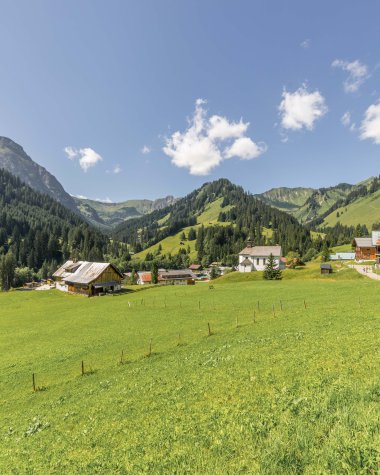 Bild eines grünen Hügels mit mehreren Häusern und Bergen im Hintergrund bei sonnigem Wetter. | © Kleinwalsertal Tourismus eGen • Fotograf: Steffen Berschin 