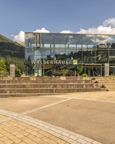 Modernes Glasgebäude des Walserhauses mit mehreren Treppen, umgeben von grünen Bergen und Bäumen, bei sonnigem Wetter. | © Kleinwalsertal Tourismus eGen • Fotograf: Steffen Berschin 