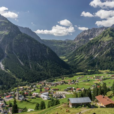 Panoramaaufnahme des Bergdorfes Mittelberg in den Alpen mit grünen Hügeln, Bergen im Hintergrund und einer blauen wolkenverhangenen Himmel. | © Kleinwalsertal Tourismus eGen • Fotograf: Steffen Berschin 