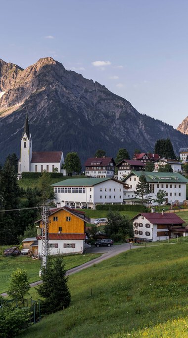 Panoramic view of a small village in the mountains with a church, surrounded by green meadows and trees, against a dramatic mountain backdrop. | © Kleinwalsertal Tourismus eGen • Photographer: Steffen Berschin 