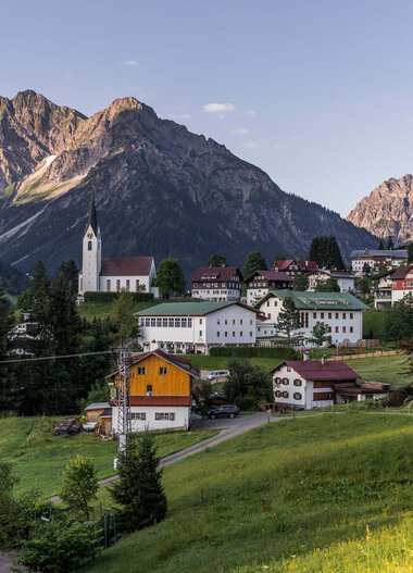 Panoramaaufnahme eines kleinen Dorfes in den Bergen mit Kirche, umgeben von grünen Wiesen und Bäumen, vor einer dramatischen Bergkulisse. | © Kleinwalsertal Tourismus eGen • Fotograf: Steffen Berschin 
