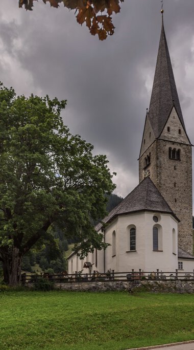 A church building with a tall spire, surrounded by trees and a house. | © Kleinwalsertal Tourismus eGen • Photographer: Steffen Berschin 