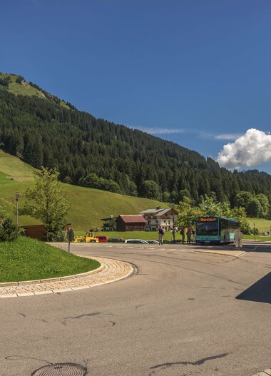 Bergdorf mit Busparkplatz inmitten grüner Hügel und Wald, schönes Sommerwetter, Bergmassiv im Hintergrund. | ©  Kleinwalsertal Tourismus eGen • Fotograf: Steffen Berschin 