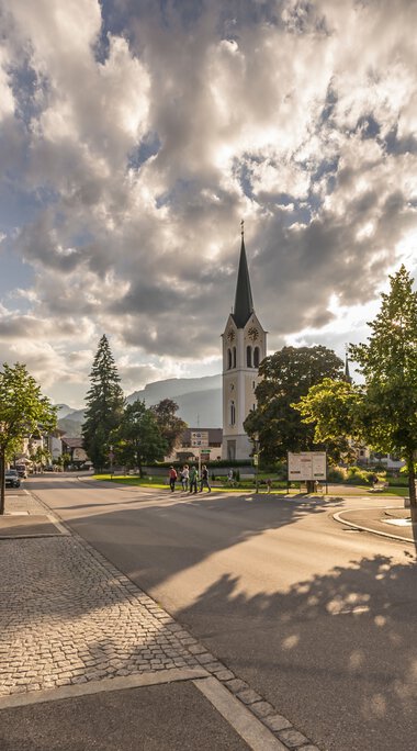 Stadtansicht mit Kirche und SonnenSchatten auf der Straße bei bewölktem Himmel, in Riezlern. | © Kleinwalsertal Tourismus eGen • Fotograf: Stefan Klauser 