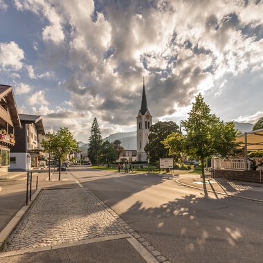Stadtansicht mit Kirche und SonnenSchatten auf der Straße bei bewölktem Himmel, in Riezlern. | © Kleinwalsertal Tourismus eGen • Fotograf: Stefan Klauser 