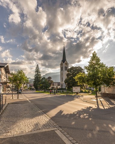 Stadtansicht mit Kirche und SonnenSchatten auf der Straße bei bewölktem Himmel, in Riezlern. | © Kleinwalsertal Tourismus eGen • Fotograf: Stefan Klauser 