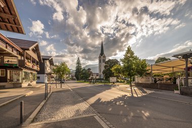 Stadtansicht mit Kirche und SonnenSchatten auf der Straße bei bewölktem Himmel, in Riezlern. | © Kleinwalsertal Tourismus eGen • Fotograf: Stefan Klauser 