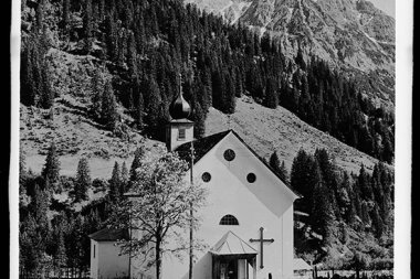 Schwarz-Weiß-Foto einer kleinen Kirche mit Kreuz, umgeben von Bäumen und Bergen, mit Wolfgänger im Hintergrund. | © Kleinwalsertal Tourismus eGen • Karl Marx Kessler Archiv