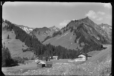 Historisches Schwarz-Weiß-Foto von einem Bergdorf mit mehreren Häusern, umgeben von Wiesen und Wäldern, mit Bergen im Hintergrund. | © Kleinwalsertal Tourismus eGen • Karl Marx Kessler Archiv