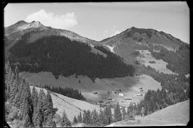 Schwarz-Weiß-Foto der Berglandschaft mit Hütten in einer talierten Landschaft, umgeben von Wäldern, mit hohen Bergen im Hintergrund. | © Kleinwalsertal Tourismus eGen • Karl Marx Kessler Archiv