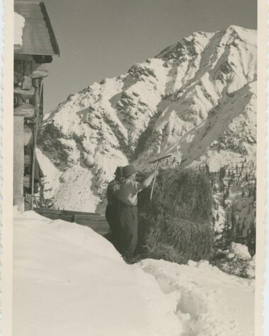 Historisches Foto einer Person beim Heugewinnung in schneereicher Berglandschaft, nahe einem Haus, im Winter. | © Kleinwalsertal Tourismus eGen • Stefan Heim