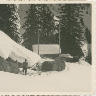 © Kleinwalsertal Tourismus eGen • Stefan Heim Historische Schwarz-Weiß-Aufnahme eines Hauses in verschneiter Landschaft, umgeben von Tannenbäumen, mit einem Menschen davor. | © Kleinwalsertal Tourismus eGen • Stefan Heim