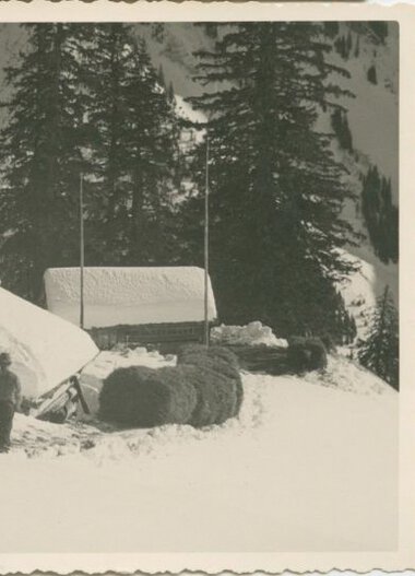Historische Schwarz-Weiß-Aufnahme eines Hauses in verschneiter Landschaft, umgeben von Tannenbäumen, mit einem Menschen davor. | © Kleinwalsertal Tourismus eGen • Stefan Heim