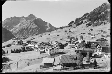 Historisches Schwarz-Weiß-Foto eines kleinen Dorfes in den österreichischen Alpen mit Kirche, umgeben von Bergen und Hütten. | © Kleinwalsertal Tourismus eGen • Karl Marx Kessler Archiv