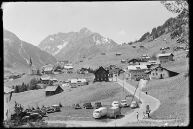Historisches Schwarz-Weiß-Foto des Ortszentrums von Hirschegg im Kleinwalsertal, mit Kirche, umliegenden Häusern, Autos und den Bergen Widderstein und Hochkalber im Hintergrund. | © Kleinwalsertal Tourismus eGen • Karl Marx Kessler Archiv