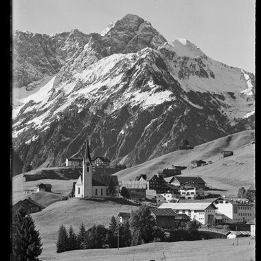 Schöne Berglandschaft im Kleinwalsertal mit Kirche und umliegenden Gebäuden, vor beeindruckenden schneebedeckten Gipfeln der Alpen. | © Kleinwalsertal Tourismus eGen • Karl Max Kessler Archiv