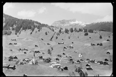 Historische Schwarz-Weiß-Fotoaufnahme von Hirschegg mit verschneiten Ifen, umgeben von Grünflächen, vereinzelten Häusern und einer Kirche. | © Kleinwalsertal Tourismus eGen • Karl Marx Kessler Archiv