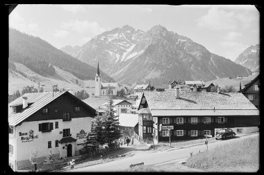 Schwarz-Weiß-Foto eines kleinen österreichischen Dorfes vor majestätischen Bergen, mit einer Kirche, traditionellen Häusern und Autos auf der Straße. | © Kleinwalsertal Tourismus eGen • Karl Marx Kessler Archiv