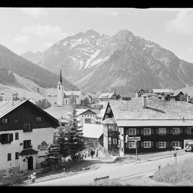 Schwarz-Weiß-Foto eines kleinen österreichischen Dorfes vor majestätischen Bergen, mit einer Kirche, traditionellen Häusern und Autos auf der Straße. | © Kleinwalsertal Tourismus eGen • Karl Max Kessler Archiv