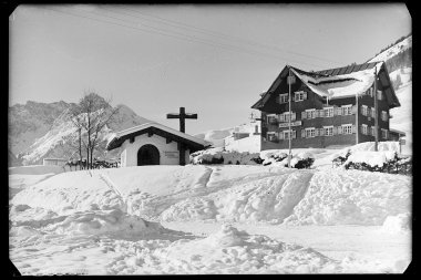Schnee bedeckter Ort mit Kapelle und Haus in den Bergen, im Winter, historische Fotografie. | © Kleinwalsertal Tourismus eGen • Karl Marx Kessler Archiv