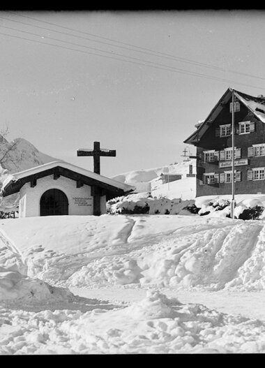 Schnee bedeckter Ort mit Kapelle und Haus in den Bergen, im Winter, historische Fotografie. | © Kleinwalsertal Tourismus eGen • Karl Marx Kessler Archiv