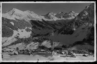 Historisches Foto des Ortzentrums von Mittelberg mit Kirche und drei Schafsalpköpfen vor verschneiten Bergen. | © Kleinwalsertal Tourismus eGen • Karl Marx Kessler Archiv