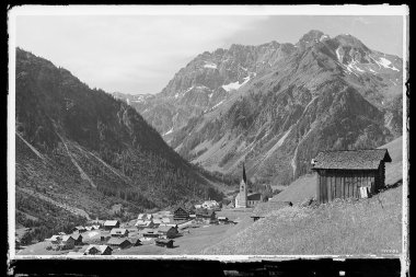 Historisches Schwarz-Weiß-Foto des Ortes Mittelberg im Kleinwalsertal, umgeben von Bergen, mit Häusern und einer Kirche im Zentrum. | © Kleinwalsertal Tourismus eGen • Karl Marx Kessler Archiv