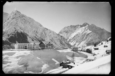 Schneebedeckte Landschaft im Mittelberg, mit Bergen im Hintergrund, einige Gebäude im Vordergrund. | © Kleinwalsertal Tourismus eGen • Karl Marx Kessler Archiv