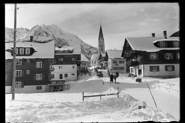 Schneebedeutes Mittelberg mit Kirche im Hintergrund, umgeben von Bergen, historische Schwarz-Weiß-Fotografie. | © Kleinwalsertal Tourismus eGen • Karl Marx Kessler Archiv