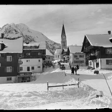 Schneebedeutes Mittelberg mit Kirche im Hintergrund, umgeben von Bergen, historische Schwarz-Weiß-Fotografie. | © Kleinwalsertal Tourismus eGen • Karl Max Kessler Archiv