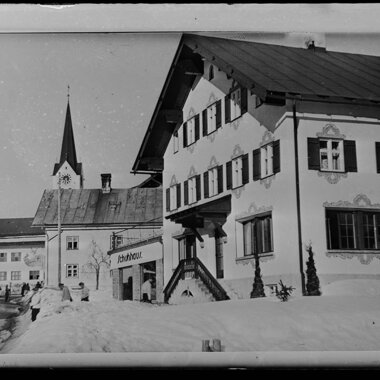 Historisches Schwarz-Weiß-Foto eines Schuh- und Modehauses im Zentrum von Riezlern, mit einem Mann und einer Frau vor dem Geschäft, im Schnee. | © Kleinwalsertal Tourismus eGen • Karl Max Kessler Archiv