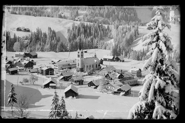 Historisches Foto des verschneiten Riezlern mit Kirche im Zentrum, umgeben von Bergen. | © Kleinwalsertal Tourismus eGen • Karl Marx Kessler Archiv
