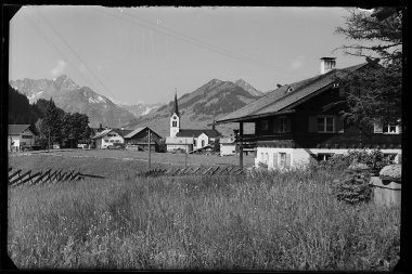 Historisches Bild von Riezlern mit Blick auf die Kirche und die umliegenden Berge, aufgenommen aus einer Wiese. | © Kleinwalsertal Tourismus eGen • Karl Marx Kessler Archiv
