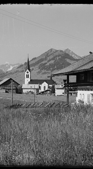 Historisches Bild von Riezlern mit Blick auf die Kirche und die umliegenden Berge, aufgenommen aus einer Wiese. | © Kleinwalsertal Tourismus eGen • Karl Max Kessler Archiv