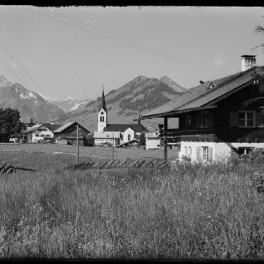 Historisches Bild von Riezlern mit Blick auf die Kirche und die umliegenden Berge, aufgenommen aus einer Wiese. | © Kleinwalsertal Tourismus eGen • Karl Max Kessler Archiv