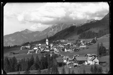 Schwarz-Weiß-Foto des Dorfes Riezlern in den Bergen, mit vielen Häusern, einer Kirche im Zentrum und umliegenden Wiesen und Bergen | © Kleinwalsertal Tourismus eGen • Karl Marx Kessler Archiv