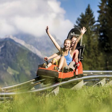 Zwei Mädchen lachen und jubeln, während sie eine Sommerrodelbahn in den Alpen hinunterfahren, umgeben von grünen Wiesen und Bergen. | © Kleinwalsertal Tourismus eGen • Oberstdorf Kleinwalsertal Bergbahnen • Fotograf: Bastian Morell