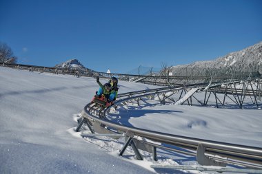 Kinder auf einer Alpenachterbahn im Schnee, umgeben von verschneiten Bergen bei klarem Himmel. | © Kleinwalsertal Tourismus eGen • Oberstdorf Kleinwalsertal Bergbahnen