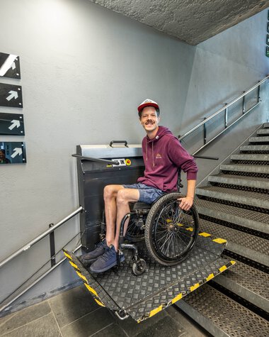 Mensch im Rollstuhl nutzt eine barrierefreie Plattform am Bahnhof mit Treppe im Hintergrund. | © Oberstdorf · Kleinwalsertal Bergbahnen Fotograf: Dominik Berchthold