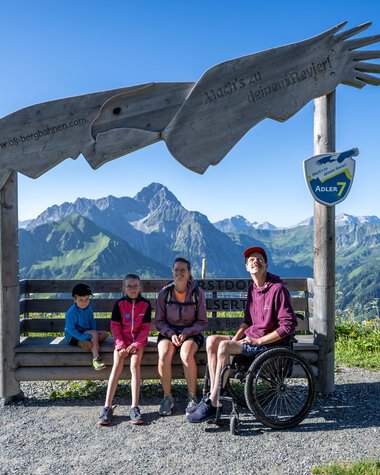 Zwei Kinder und zwei Erwachsene, einer mit Rollstuhl, sitzen auf einer Bank vor einer großen Skulptur eines gebenden Falken mit Bergpanorama im Hintergrund, bei den OK-Bergbahnen in Oberstdorf. | © Oberstdorf · Kleinwalsertal Bergbahnen Fotograf: Dominik Berchthold