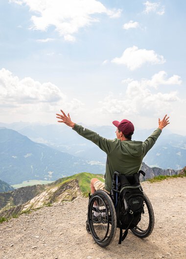 Eine Person im Rollstuhl mit offenem Arm in einer Berglandschaft, Blick auf Berge und Himmel, zeigt Freude und Freiheit. | © Oberstdorf · Kleinwalsertal Bergbahnen Fotograf: Frithjof Kjer