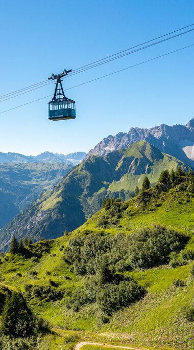Bergbahn fährt über grüne Hänge und Berge im Kleinwalsertal, mit einem klaren blauen Himmel im Hintergrund. | © Oberstdorf · Kleinwalsertal Bergbahnen