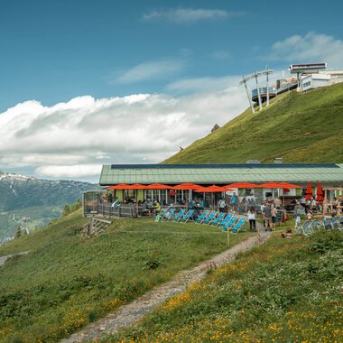 Bergrestaurant mit Terrasse auf einer grünen Berghänge im Alpengebiet, Blick auf die Berge im Hintergrund. | © Oberstdorf · Kleinwalsertal Bergbahnen Fotograf: Basti Heckl