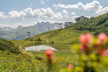 Berglandschaft mit Fellhornbahn über einer kleinen Wasserfläche, grünen Hügeln und Bergketten im Hintergrund. | © Oberstdorf · Kleinwalsertal Bergbahnen Fotograf: Basti Heckl